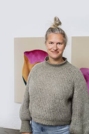 Eine Person mit einem grauen Pullover und hochgestecktem Haar lächelt in die Kamera, im Hintergrund sind abstrakte Gemälde.