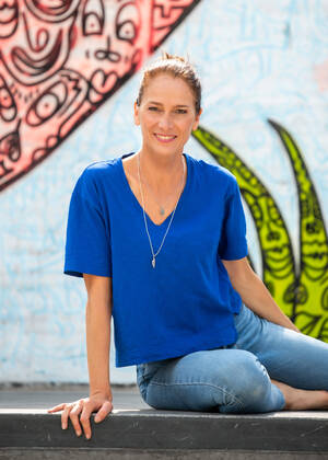 Eine Frau in einem blauen T-Shirt sitzt lächelnd vor einer farbenfrohen, bemalten Wand.