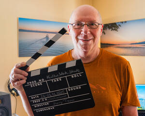 Ein Mann in einem orangefarbenen T-Shirt hält eine Filmklappe in der Hand und lächelt in die Kamera, während im Hintergrund ein Landschaftsbild an der Wand hängt.