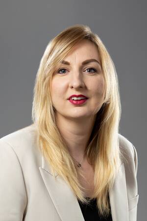 Eine Frau mit langen blonden Haaren trägt einen cremefarbenen Blazer und schaut in die Kamera.
