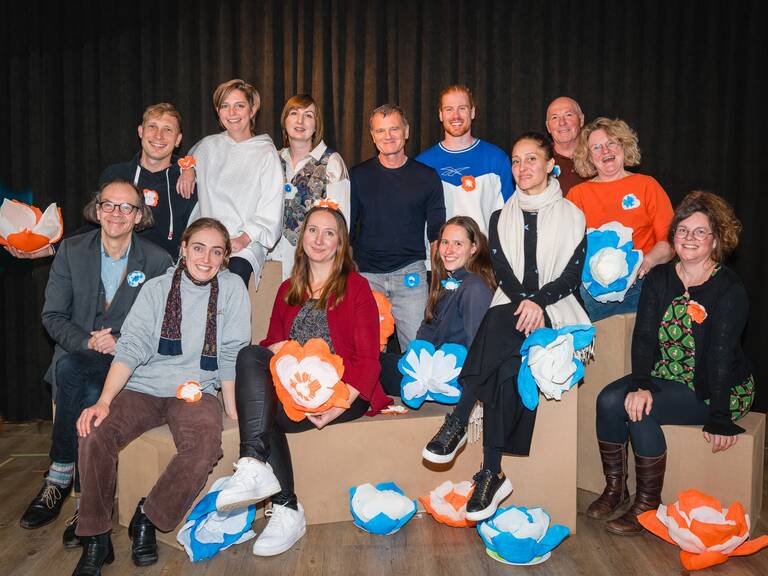 Eine Gruppe von dreizehn lächelnden Menschen posiert auf einer Bühne vor einem schwarzen Vorhang, umgeben von großen, farbenfrohen Papierblumen in Orange, Blau und Weiß.