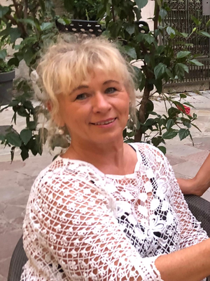 Eine Frau mit blonden Haaren sitzt lächelnd in einem Stuhl, umgeben von grünen Pflanzen.