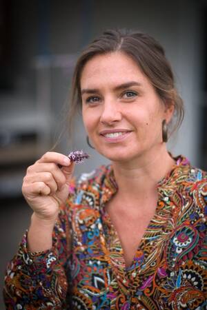 Eine lächelnde Frau mit langen Haaren hält ein kleines lila Blümchen und trägt eine bunte, gemusterte Bluse.