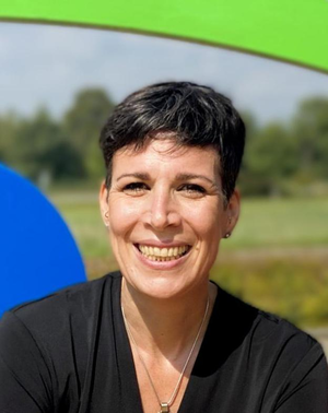 Eine lächelnde Frau mit kurzen Haaren trägt ein schwarzes Oberteil und steht vor einem bunten, unscharfen Hintergrund im Freien.