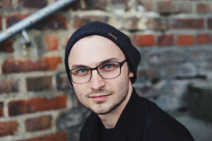 Ein Mann mit Brille und Mütze schaut vor einer unscharfen Backsteinmauer in die Kamera.