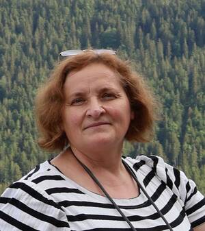 Eine Frau mit schulterlangem, braunem Haar und gestreiftem Oberteil steht vor einem Hintergrund aus Bäumen.