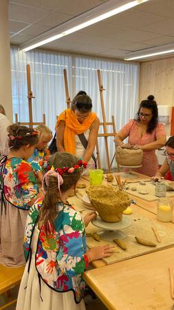 In einem Kunsthandwerksraum formen mehrere Erwachsene und Kinder in bunten Bekleidungen gemeinsam mit Lehm an einem Tisch.