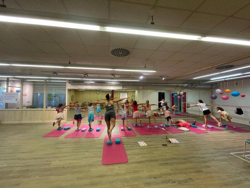 Mehrere Kinder üben eine Yoga- oder Tanzübung auf rosa Matten in einem Raum mit Holzboden und farbigen Bällen an der Wand.