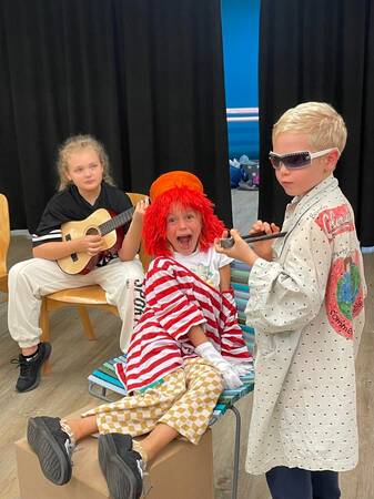 Drei Kinder sind in einem Raum; eines spielt Gitarre, ein anderes trägt ein buntes Kostüm, und das dritte Kind im weißen Mantel trägt eine Sonnenbrille und hält ein Mikrofon.