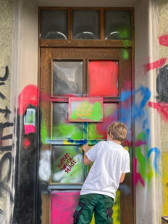 Ein Kind in einem weißen T-Shirt bemalt eine Holztür mit buntem Graffiti, auf der "Kultur Werk Stadt" steht.