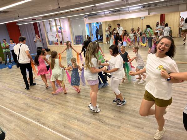 In einem Tanzstudio tanzen Erwachsene und Kinder gemeinsam in einem Kreis, während einige zuschauen und fotografieren.