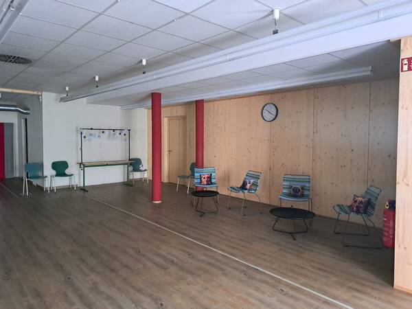 Ein heller Raum mit Holzwänden und -boden zeigt eine Sitzgruppe aus bunten Stühlen und Tischen, einem Whiteboard und einer Wanduhr.