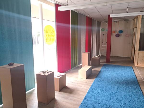 Ein Raum mit farbigen Vorhängen, hölzernen Sockeln und einem blauen Teppich, dekoriert mit bunten Kugeln an der Wand.