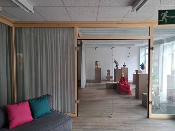 Ein Raum mit Glaswand zeigt eine Ausstellung mit verschiedenen Skulpturen auf Sockeln, darunter eine in rotem Stoff gehüllt.