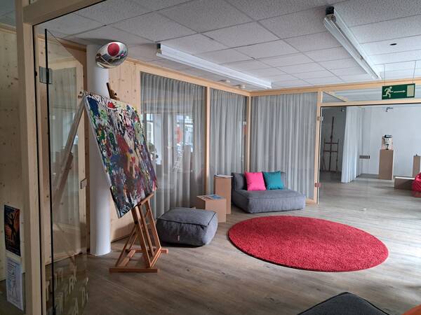 Ein modern eingerichteter Raum mit einem großen abstrakten Gemälde auf einer Staffelei, einem runden roten Teppich, bequemen Sitzmöglichkeiten und Kissen in leuchtenden Farben.