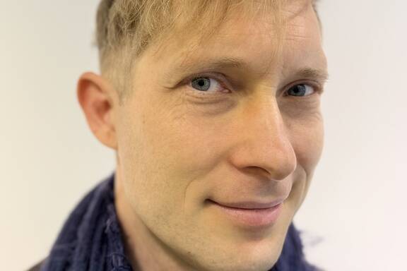 Eine lächelnde Person mit blonden Haaren trägt einen dunkelblauen Schal und blickt leicht seitlich.